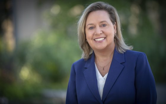 A headshot of a person with shoulder length dirty blond hair, in a dark blue jacket with white blouse, smiling at the camera.